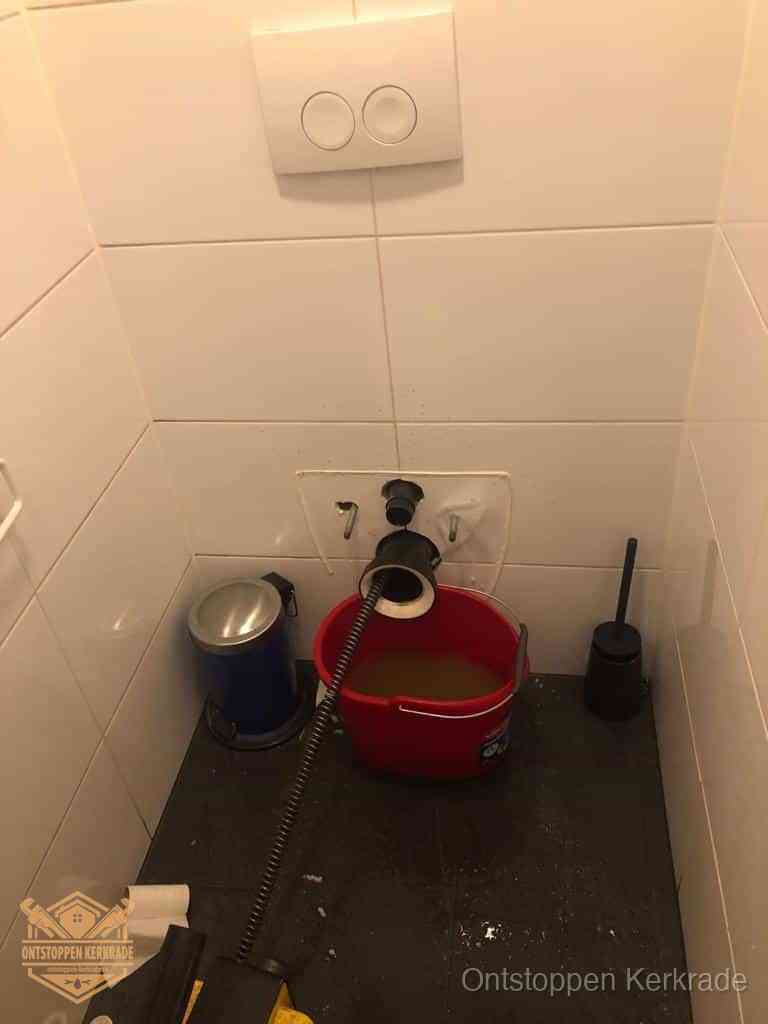 Een ontstopte wand-wc met verwijderde toiletpot waar een ontstoppingsveer in de wanduitgang verdwijnt, het achterhalen waar de verstopping zit in Kerkrade