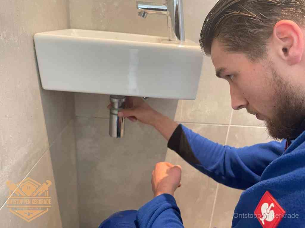 Een monteur draait met de hand de zwanenhalssifon onder een fonteinwasbak in de toiletruimte los om de bron van rioollucht in Kerkrade te vinden