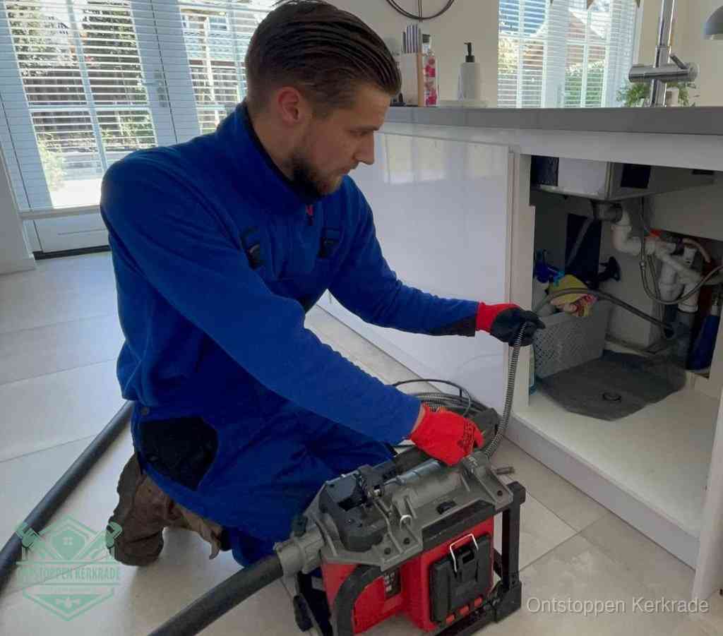 Een monteur met rode handschoenen knielt in een keuken en voert een elektrische ontstoppingsveer in tijdens het professioneel afvoer ontstoppen in Kerkrade
